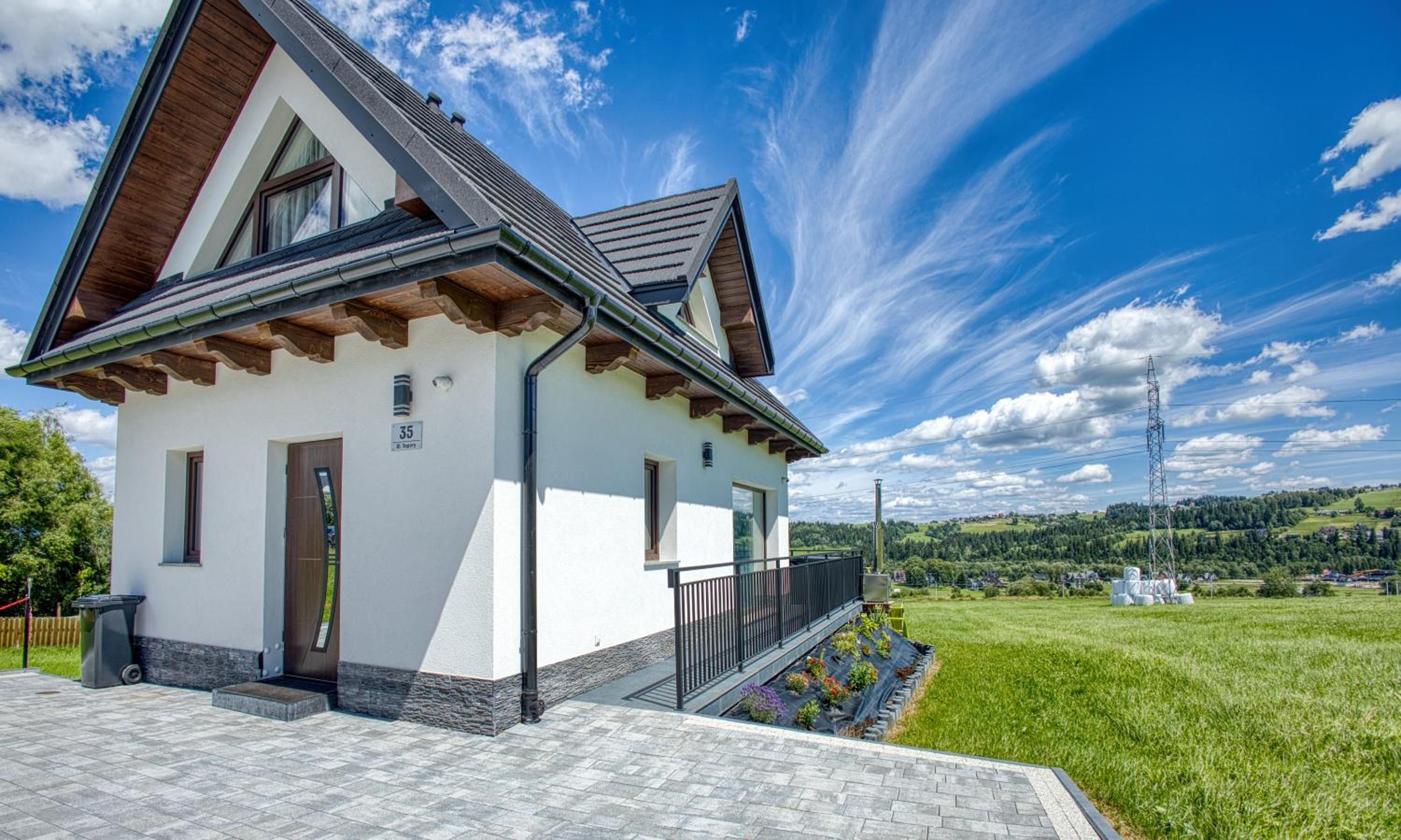 Biały dom Mery House z ciemnym dachem i drewnianymi belkami, obok zielona łąka i wzgórza pod błękitnym niebem