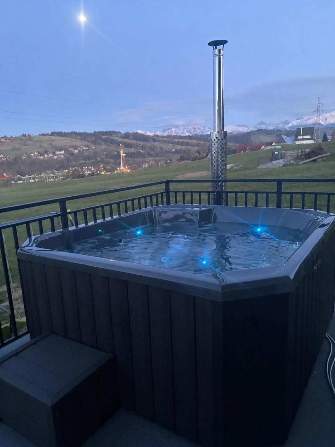 Podświetlone jacuzzi przy balustradzie, w tle łąka i ośnieżone góry o zmierzchu